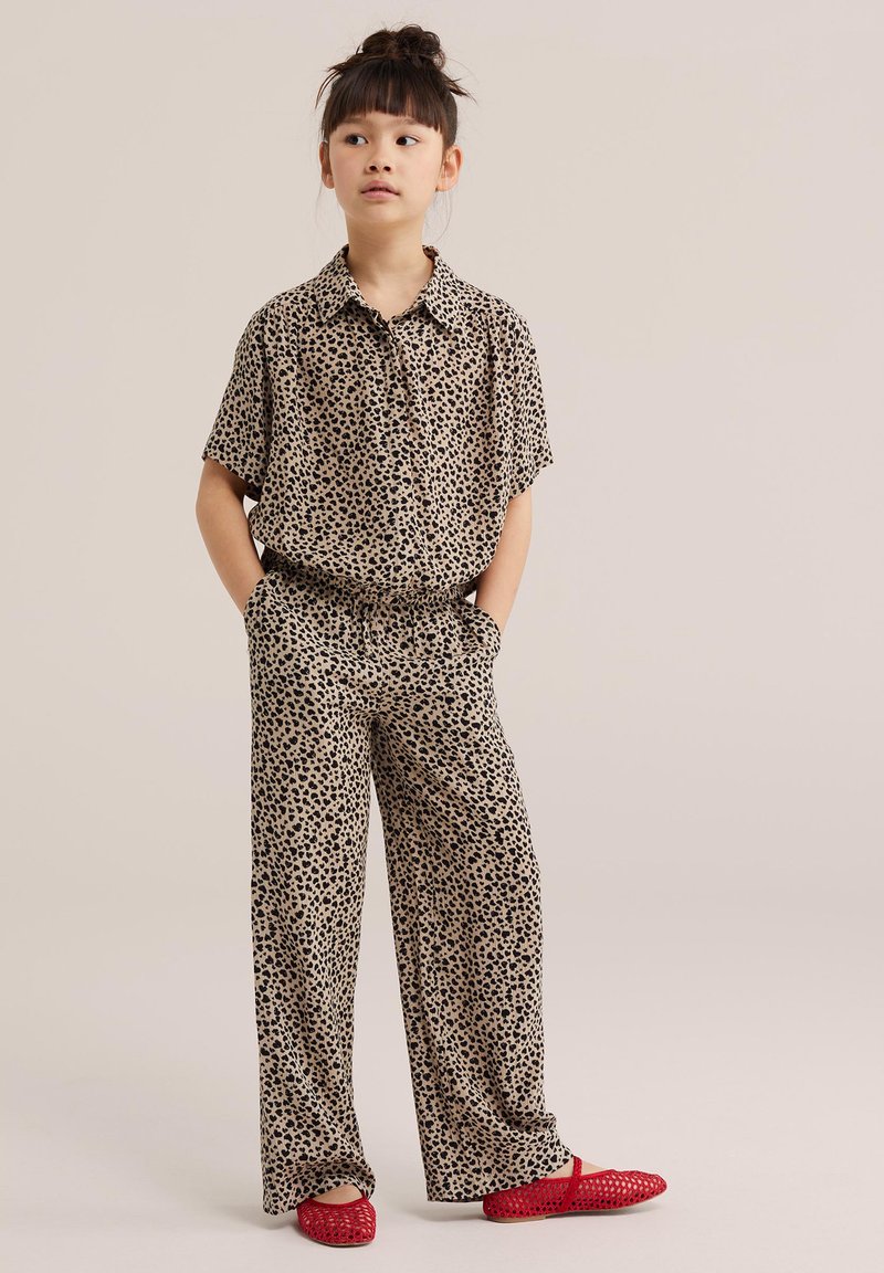 Młoda dziewczyna z koczkiem, ubrana w beżową koszulę na guziki z krótkim rękawem oraz pasujące szerokie spodnie w czarne kropki, czerwone wsuwane buty z plecionki.