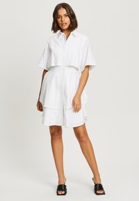 Robe blanche avec un col, des manches courtes en forme de cape, un ourlet superposé et une fermeture éclair à l'avant. Portée avec des sandales noires à glissière.