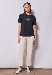 T-shirt en coton noir avec texte blanc, pantalon large crème, et baskets beiges avec accents noirs, le tout sur un fond neutre.