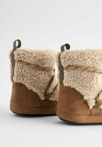 Tanfarbene Slipper mit softem, flauschigem, beigem Schaffell-Obermaterial, ausgestattet mit olivgrünen Riemen und strukturierten braunen Sohlen für besseren Grip.