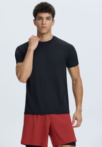 Camiseta deportiva negra de manga corta con cuello redondo y textura suave, combinada con pantalones cortos rojos. Diseño ligero adecuado para ropa deportiva.