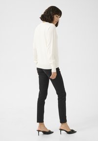 Weißer Strickpullover mit langen Ärmeln, kombiniert mit eng anliegenden schwarzen Jeans und offenen schwarzen High-Heel-Pantoletten.