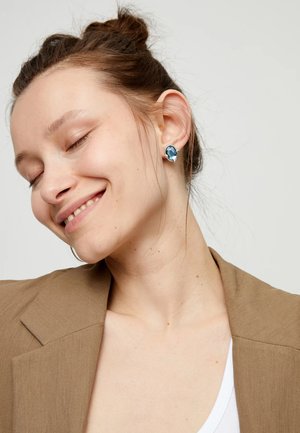 Pendientes de plata con una gran piedra azul ovalada enmarcada en un marco pulido, llevados por una modelo con un blazer de color neutro y una camiseta blanca.