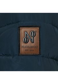 Il giubbotto imbottito blu navy presenta una toppa in pelle marrone con il testo in rilievo "NAVAHOO EST 2014" e un contorno in feltro scuro.