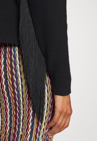 Main droite reposant contre une jupe colorée à motif ondulé, portant un haut noir texturé à manches longues avec un détail de franges noires sur le côté.
