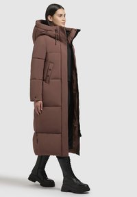 Barna, hosszú puffermellény kapucnival, steppelt mintával, fekete gombokkal és oldalsó zsebekkel. Fekete öltözék fölött viselve, térdfelett érő fekete csizmával.