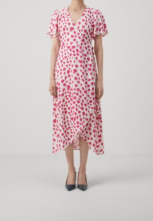 Kobieta ubrana w białą, midi sukienkę kopertową w czerwone wzory serc, z falującymi rękawami, w czarnych butach na szpic, stojąca na jednolitym tle.