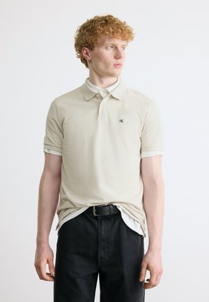 Noor mees laineliste punaste juustega, kes kannab beeži polosärki, mis on kergelt mustadesse pükstesse toppitud, vaadates paremale tavalise valge tausta ees.