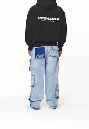 Person trägt schwarzen Hoodie mit dem Schriftzug "PEGADOR" und hellblaue Cargo-Jeans mit einem großen blauen Aufnäher mit dem Text "NEVER TOO LATE" auf der Rückseite.