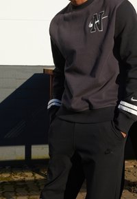 Tmavě šedá mikina s černými rukávy, na které se nachází výrazné logo 'N' a Nike Swoosh. Pruhované detaily na manžetách. Černé kalhoty s logem.