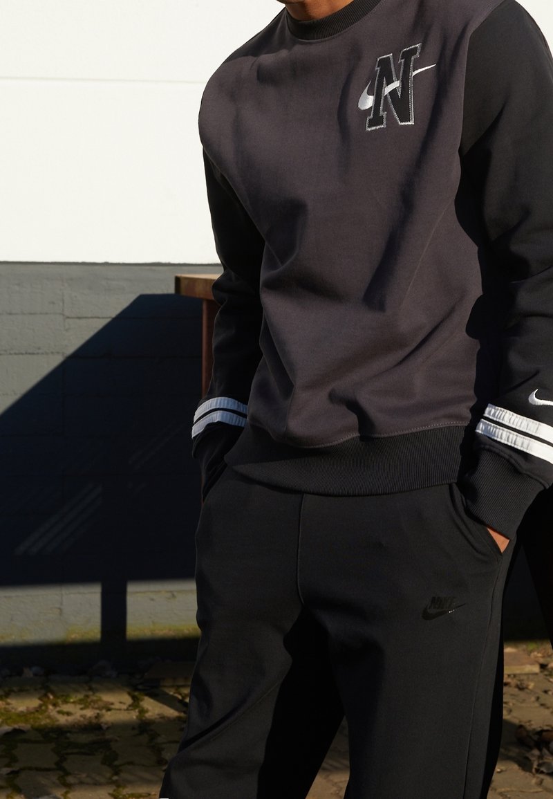 Tmavě šedá mikina s černými rukávy, na které se nachází výrazné logo 'N' a Nike Swoosh. Pruhované detaily na manžetách. Černé kalhoty s logem.