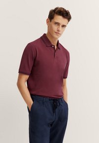 Polo di colore bordeaux con colletto classico e accentuazioni a righe blu navy sulle maniche, abbinato a pantaloni blu scuro con cordino.