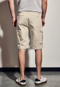 Béžové cargo šortky s bočními kapsami, délka po kolena, vyrobené z prodyšného materiálu. Nosené s bílými teniskami s černým akcentem na patě.