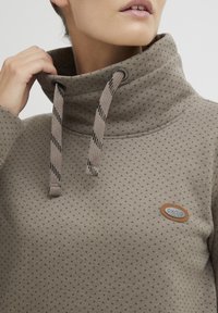 Felpa in pile marrone a motivo con colletto alto, dotata di cordini decorativi e di una piccola etichetta del marchio. Presenta punti testurizzati su tutta la superficie.