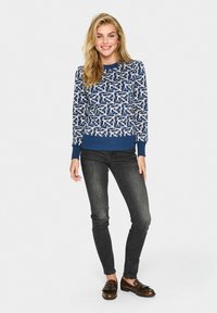 Pull bleu avec motif abstrait blanc, col rond et poignets côtelés, associé à un jean foncé et des mocassins marron.