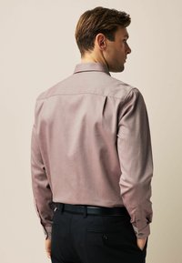 Langärmliges, burgunderfarbenes Hemd mit subtilen Fischgrätmuster und Manschettenknöpfen. Getragen mit schwarzen Hosen, die eine maßgeschneiderte Passform und glatte Textur zeigen.