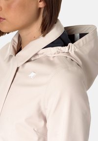Giacca beige chiaro con cappuccio aderente, caratterizzata da un esterno liscio, cuciture sottili e un piccolo logo bianco sul petto.