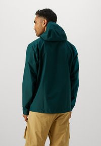 Veste imperméable vert foncé avec capuche, arborant une texture lisse et une coupe décontractée, portée sur un pantalon cargo beige.