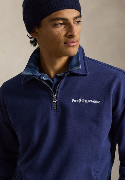 Νέος άνδρας με φακίδες που φοράει σκούφο ναυτικού χρώματος και φούτερ Polo Ralph Lauren με φερμουάρ μέχρι το στήθος σε ναυτικό χρώμα πάνω από μπλε πουκάμισο με καρό μοτίβο, κοιτάζει προς τα δεξιά.