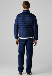 Marineblå dunjakke med quiltet tekstur, høj krave og logodetalje ved halsen, parret med mørke jeans og casual sko.