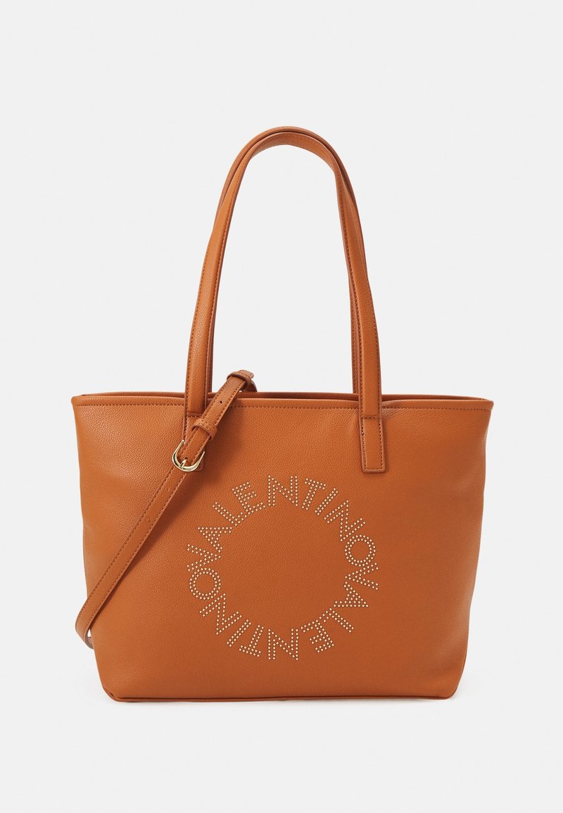 Brown leather tote bag with twin handles and an adjustable shoulder strap. Features a circular studded logo detail on the front.