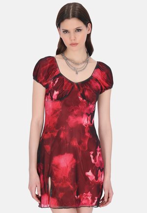 Jeune femme portant une robe courte fleurie rouge et noire avec des manches bouffantes et un collier en chaînes argentées superposées, debout devant un fond uni.