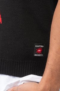 Svart strikket genser med ribbestrikket kant, med et rødt stjernemønster og et svart merke som sier "KAOTIKO SOCIETY."