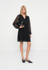 Nainen mustassa läpikuultavassa pitkähihaisessa mekossa, joka pitää mustaa teksturoidusta clutch-laukusta, pukeutuneena mustiin teräväkärkisiin slingback-korkokenkiin, seisoo valkoista taustaa vasten.