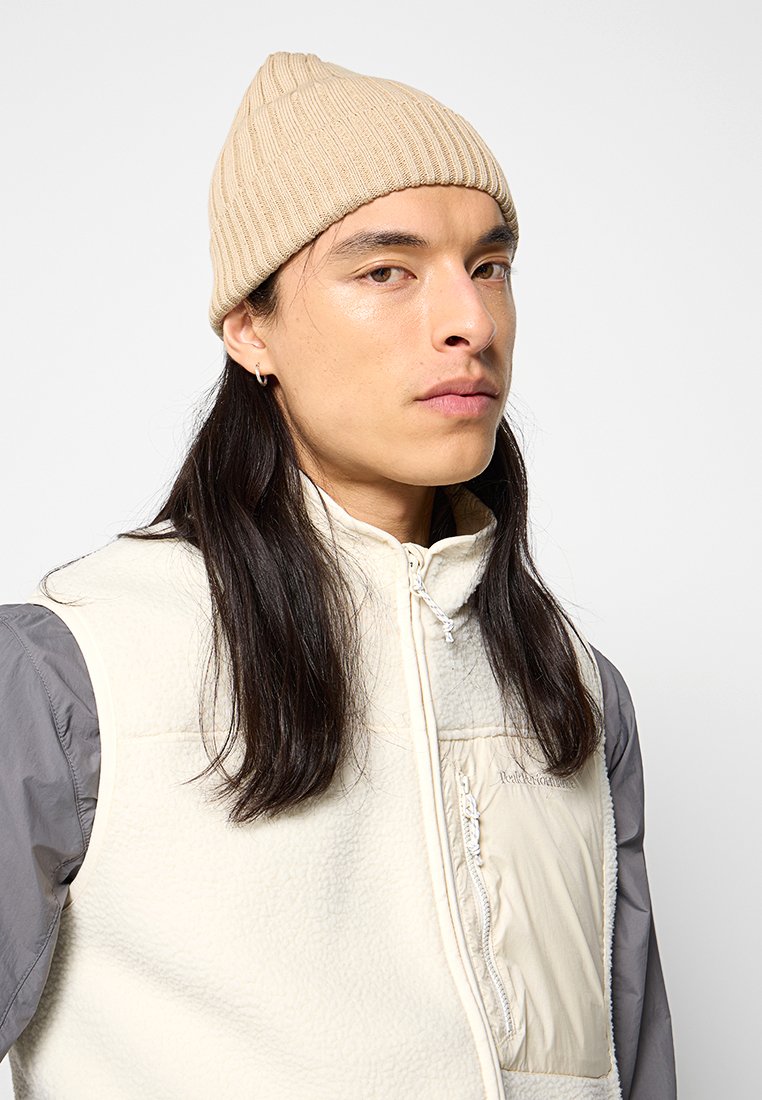 Beżowa dziana czapka beanie o prążkowanej fakturze, noszona nad czołem. W komplecie z kremową kamizelką polarową z szarymi rękawami i zapięciem na zamek.