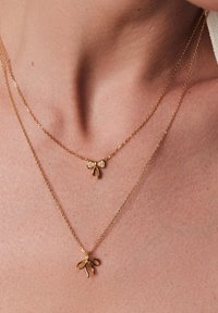 Złote naszyjniki nałożone na siebie z wisiorkami w kształcie kokard; jeden z nich ma małą kokardkę z kamieniami, a drugi większą, gładką kokardę. Delikatny wzór łańcuszka.