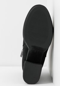 Chaussure noire avec une surface texturée et une semelle en caoutchouc. Elle présente un bout arrondi et un marquage subtil. Le design met l'accent sur l'adhérence et la stabilité.