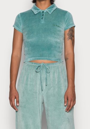 Frau trägt ein türkisfarbenes, kurz geschnittenes Velours-Poloshirt und passende Hosen mit Kordelzug, mit floralen Tattoos am rechten Arm.