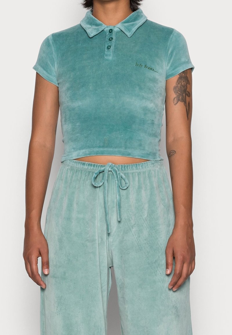 Femme portant un polo court en velours bleu sarcelle et un pantalon à cordon assorti, avec des tatouages floraux sur le bras droit.