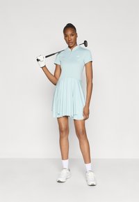 Svijetloplavi golf haljina s kratkim rukavima, zip ovratnikom i lepršavom suknjom. Model drži golf palicu, noseći bijelu rukavicu i sportske cipele.