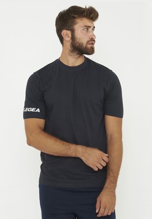 Man met baard die een effen zwart T-shirt met korte mouwen draagt met de tekst "LEGEA" op de linker mouw, kijkt naar rechts tegen een witte achtergrond.