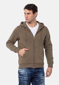 Olijfgroene zip-up hoodie gemaakt van zachte stof, met een voorzak en verstelbare trekkoordkap. Draagt met blauwe denim jeans.