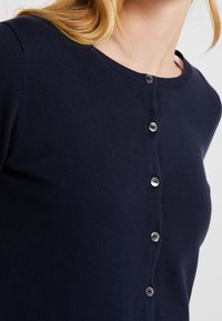 Navyblauer Cardigan aus weichem Material, mit einem runden Ausschnitt und sechs silbernen Knöpfen vorne. Glatte Textur mit figurnahem Design.
