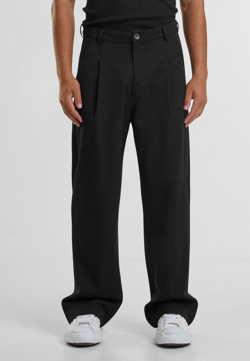 Pantalons plissés noirs à coupe décontractée, en tissu doux, avec poches latérales et fermeture à bouton. Assortis avec des baskets blanches.