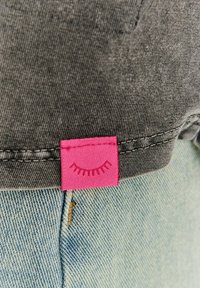 Šedá tkanina s prošitým lemem nad modrými džíny z denimu, na kterých je malá růžová visačka s motivem zavřených řas.