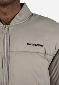 Gaiši pelēks pūkains mētelis ar ribotu apkakli, ar rāvējslēdzēju un melnu "PEGADOR" logo uz krūtīm. Gluda materiāla struktūra ar quilted sekcijām.