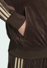 Brown velvet tracksuit top featuring a zippered pocket, textured fabric, and contrasting light yellow three-stripe design along the sleeves.