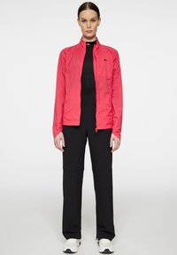 Lätt, ljusrosa jacka med dragkedja fram, elastiska ärmar och logotypdetalj. Buren över en svart outfit med utsvängda byxor och vita sneakers.