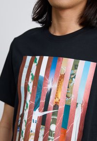 En person med langt hår viser en sort T-shirt med et levende, flerpanel grafisk design med forskellige farverige billeder og et Nike-logo.