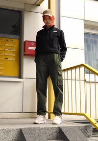 Must musta; musta fliisikombinesoon, oliivrohelised cargo-püksid taskutega, beežid tennisekingad valgete detailidega ja beež müts. Seisab treppidel.