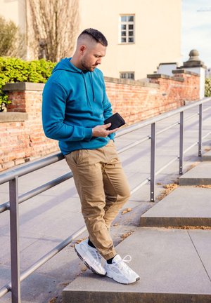Blue hoodie, beige cargo pants, and white sneakers. Man leans against a railing, holding a black smartphone, with brick wall in background.