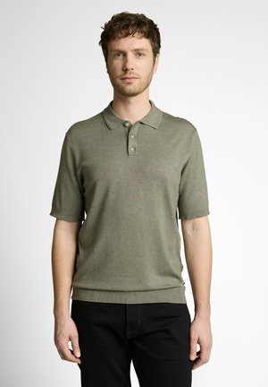 Homme aux cheveux bruns bouclés portant un polo à manches courtes vert olive et un pantalon noir, debout devant un fond blanc uni.