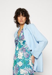 Blazer azul claro sobre um vestido com estampa floral apresentando tons de turquesa, roxo e verde. O blazer tem um corte descontraído e detalhes com botões.