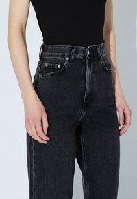 Persona che indossa jeans neri a vita alta e un top senza maniche nero infilato, con le mani rilassate lungo i fianchi.