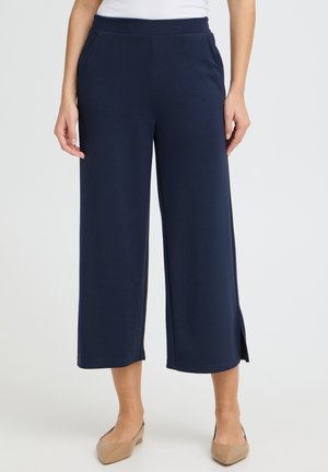 Kobieta nosząca granatowe, szerokie spodnie o długości 7/8 z bocznymi rozcięciami oraz beżowe buty na niskim, blokowym obcasie.