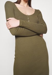 Kvinna med armarna i kors som bär en olivgrön, långärmad, ribbad klänning med tre små knappar på bröstet och en rundad halsringning.